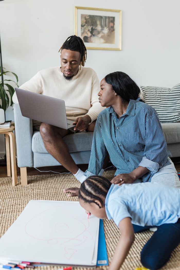 Parents engage with child drawing while using a laptop in a cozy living room setting.